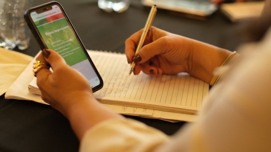 Mãos seguram um celular com o material da aula enquanto anotam em um caderno